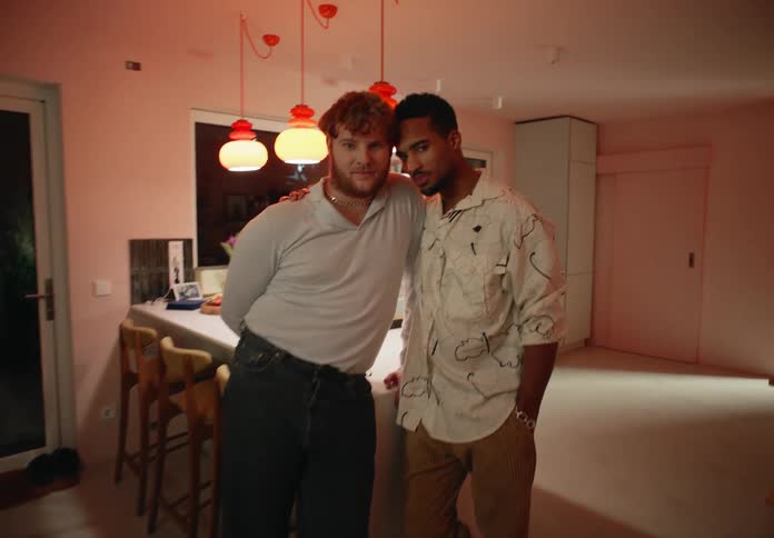 Two men stand close together posing in kitchen one touching the others neck