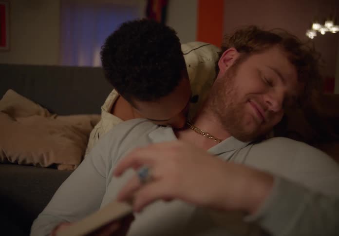 A man sits on the floor and reads as he partner lies on the couch behind him and starts to kiss him