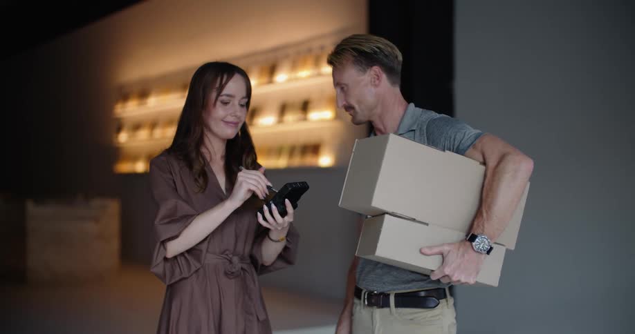A delivery man is carrying boxes into a business as he hands the signature pad to a woman and she signs for the packages