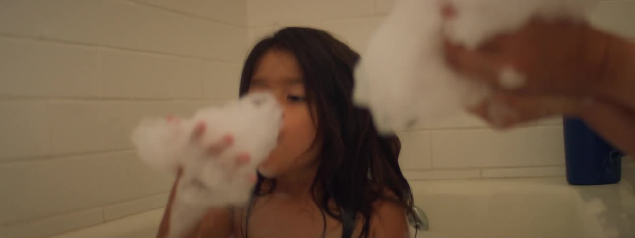 A little girl is sitting in a bubble bath blowing on the bubbles and her mother is next to her holding handfuls of bubbles