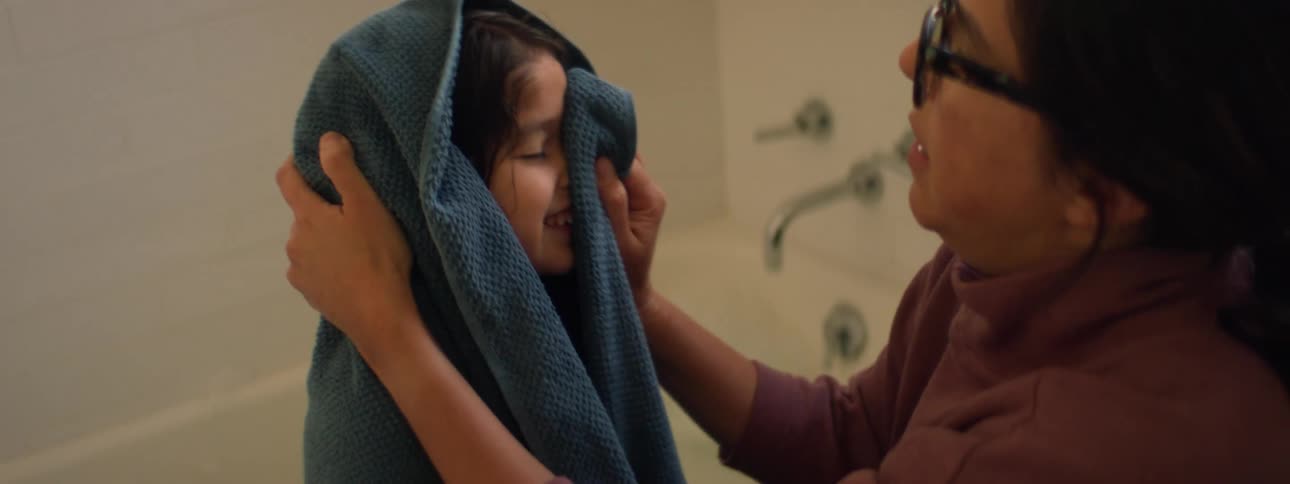 A woman is kneeling near the bathtub as her young daughter is covered with a towel and she dries her face