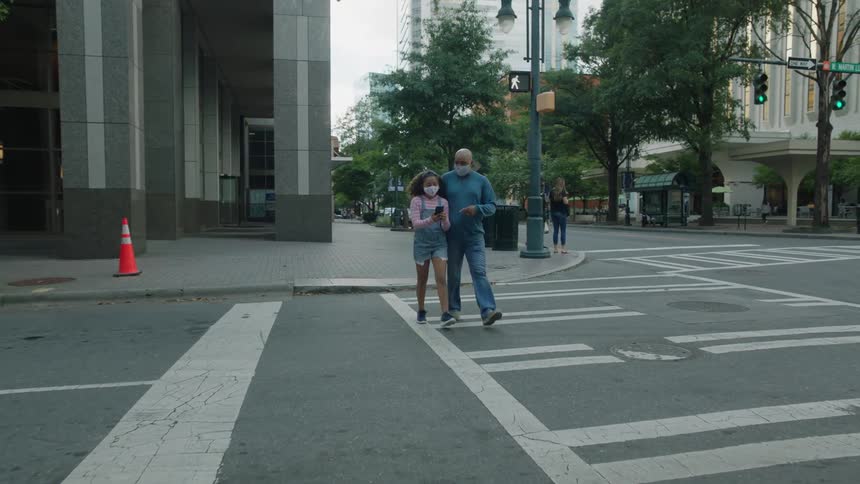 Mask wearing father and daughter use smartphone to navigate as they cross downtown street