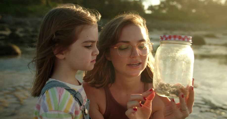 One daughter and her mother examine shells in jar as her sister plays in surf in the background