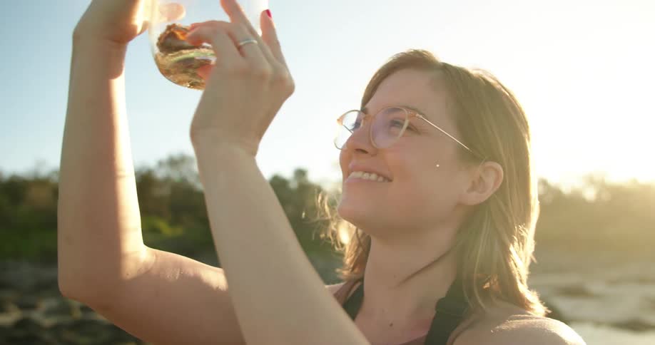 A woman is standing along the shoreline as she holds a glass jar in the air looking at the seashells inside