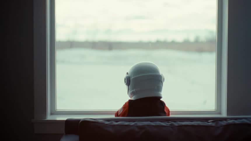 Child wearing white helmet stands looking out window at lake
