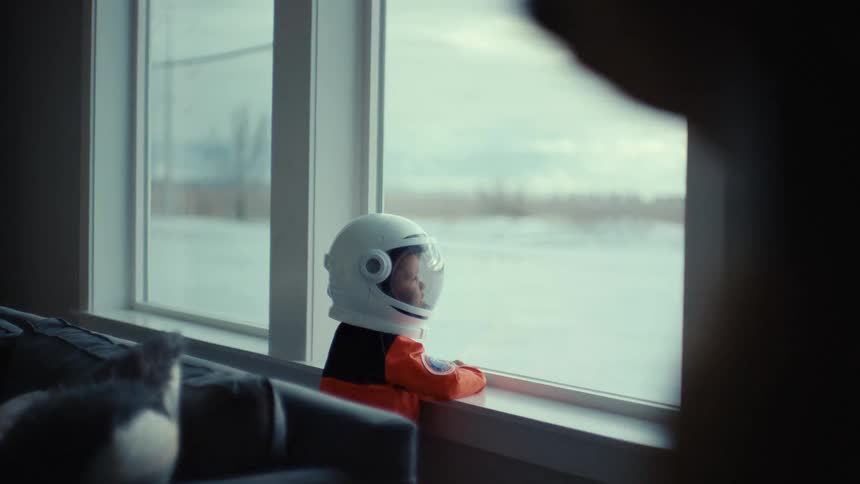 A toddler wearing a racing outfit and helmet looks out the window as an adult walks back and forth through the frame