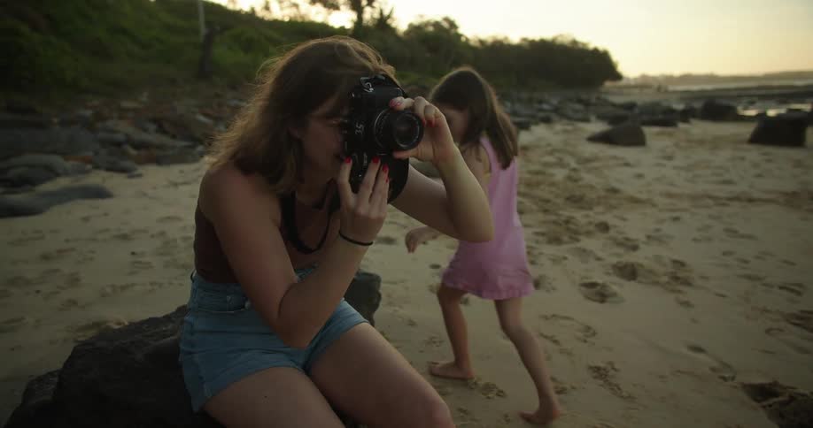 A woman is kneeling down on the beach holding her camera as she takes a picture and her two young daughters are walking on the sand to see her