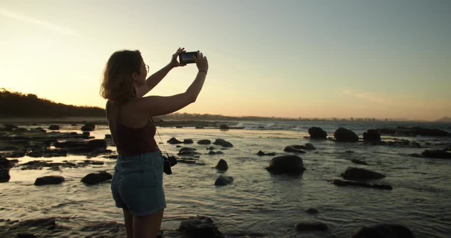 A woman is standing along the shoreline as waves roll in splashing over rocks along the coast with a camera around her neck as she holds her phone high in the air taking a picture