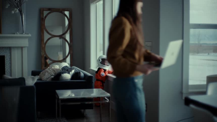 A child staring out the window in a space suit while a woman walks around on the phone carrying a laptop