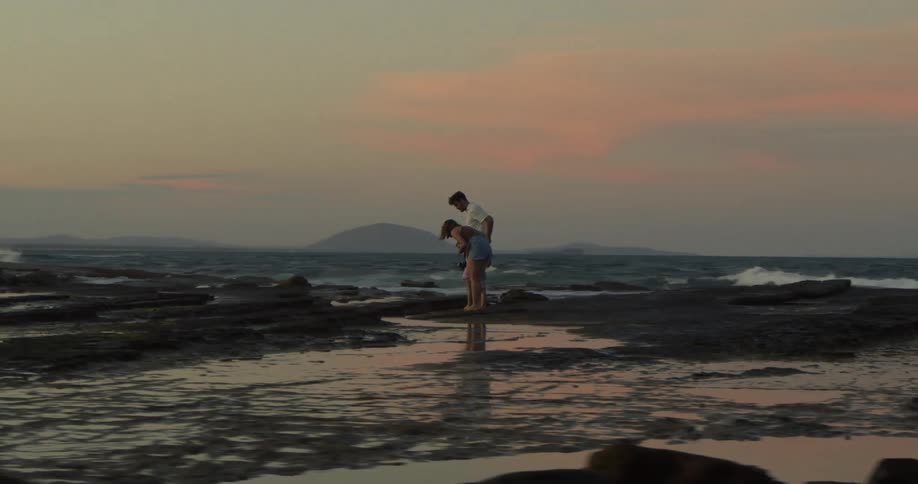 A family with two young girls are standing on the shore looking at water puddles along the coast as waves roll in