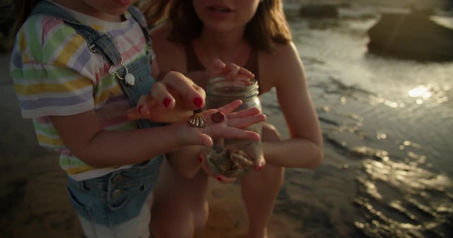 A woman is kneeling along the shoreline with her young daughter looking at seashells they are collecting in a jar as waves roll in splashing over rocks along the coast