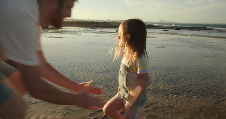 A woman is standing along the shoreline holding her camera as her young daughter runs to her father and he holds her in the air and spins her around
