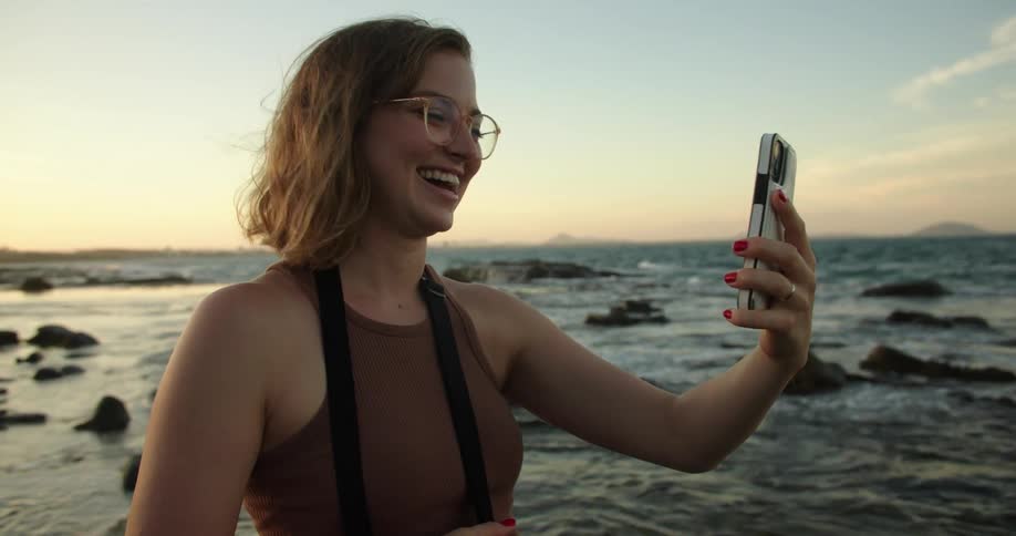 A young woman is using her phones video camera to someone a kiss with a beach coastline in the background