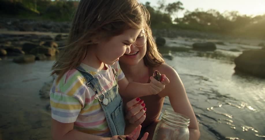 Woman holds glass jar as she and girl examine collected shells on beach on sunny day