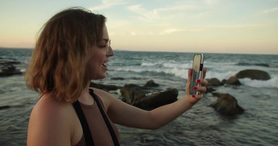 A woman waves to someone during a video call while on the beach