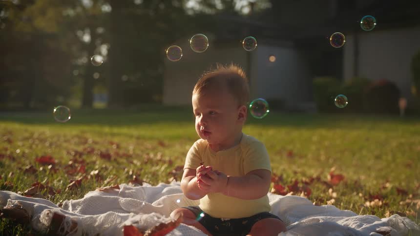 Toddler boy sits on rug on green lawn in the yard looking at bubbles floating around