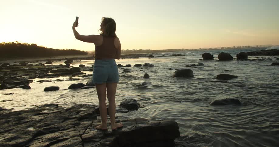 A woman is standing along the shoreline as waves roll in splashing over rocks along the coast as she holds her phone up showing herself near the water