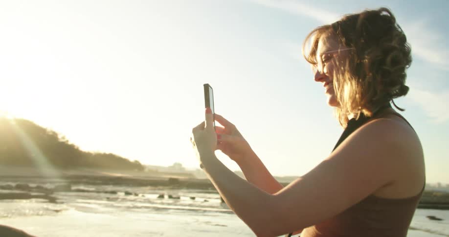 A woman is standing along the shoreline as waves roll in splashing over rocks along the coast with a camera around her neck as she holds her phone smiling and laughing