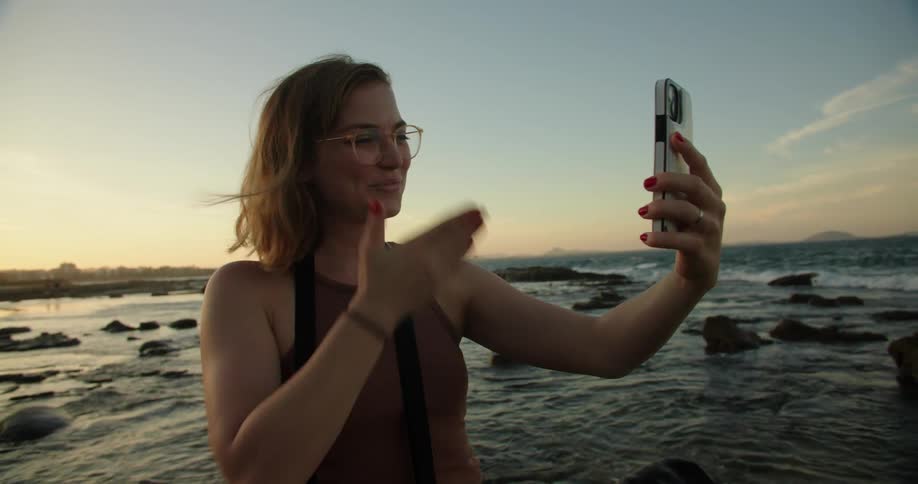 A woman is standing along the shoreline as waves roll in splashing over rocks along the coast holding out her cell phone and waving