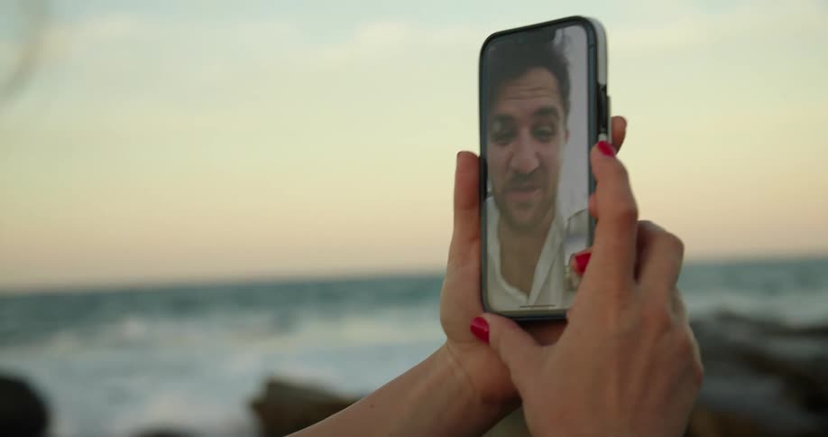 A woman is standing along the shoreline as waves roll in splashing over rocks along the coast as she holds her phone having a video conference with her husband and daughter