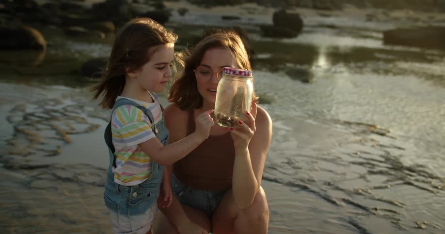 Mother and daughter on beach smile while looking at jar fool of shells