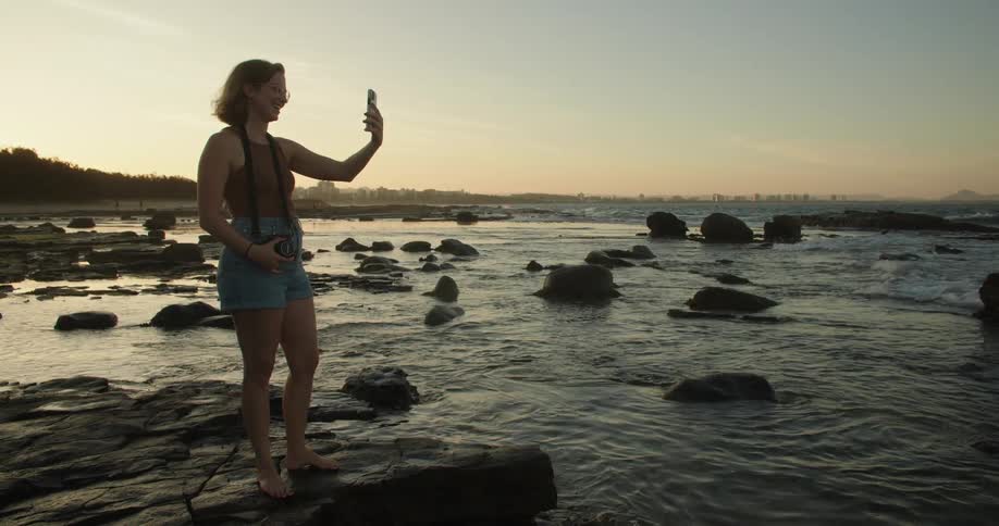 A woman is standing on rocks along the shoreline as waves roll in splashing over rocks along the coast holding out her cell phone and waving