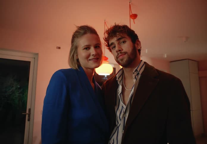 Blonde woman with blue dress and man with beard and black jacket pose for the camera in the room
