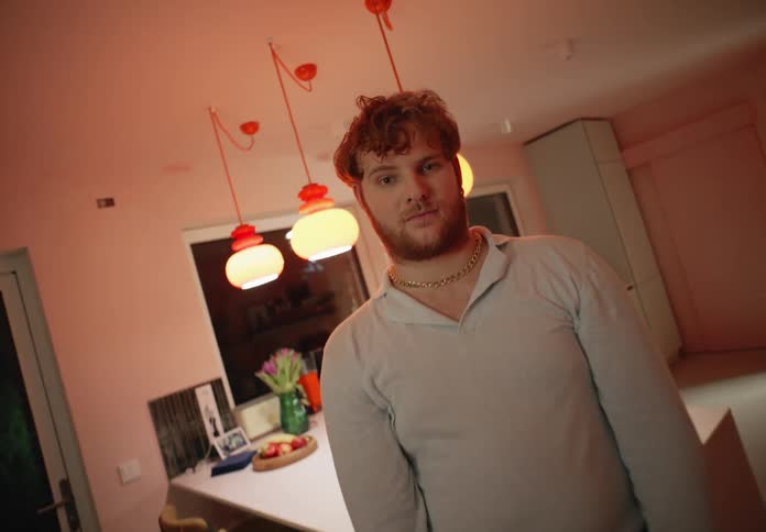 A man is standing in a room near a counter and chairs with his hands clasped in front of him