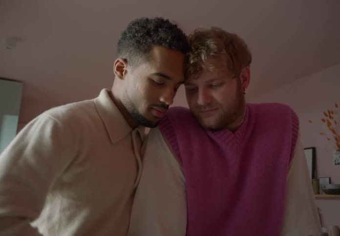 A male couple stands with their heads together in a kitchen