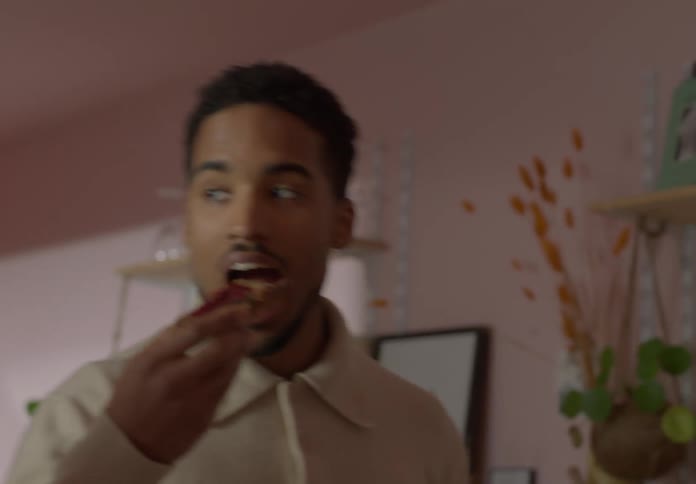 A man walks around another man and eats a piece of toast as he smiles at him