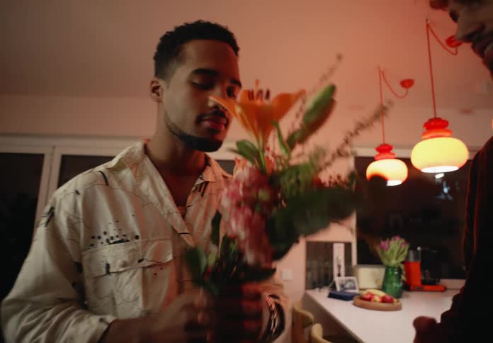 A man is standing in a room holding a bouquet of flowers and he smells them as a man standing next to him laughs and elbows his arm