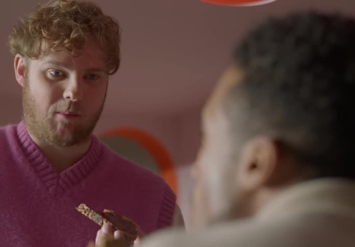 A man is eating a piece of toast as he stands talking to a man sitting down near him