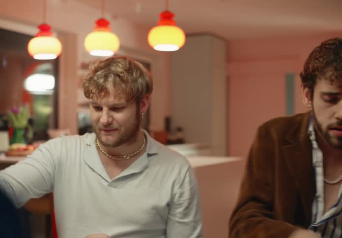 Two men sit at the dinner table eating and laughing while one steals bread off the others plate