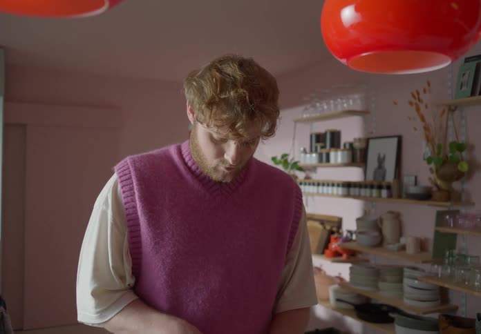 Man with beard and pink sleeveless pullover cooks while his lover enters kitchen