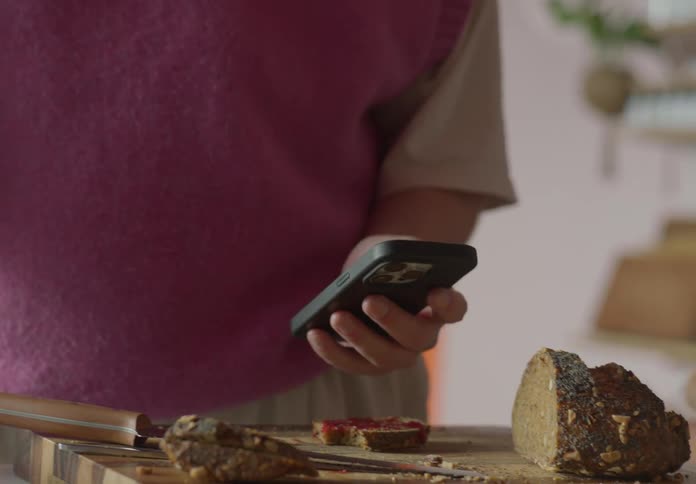 A man is standing near a table with bread on a cutting board as he pulls his phone out of his pocket and holds it out to show his friend