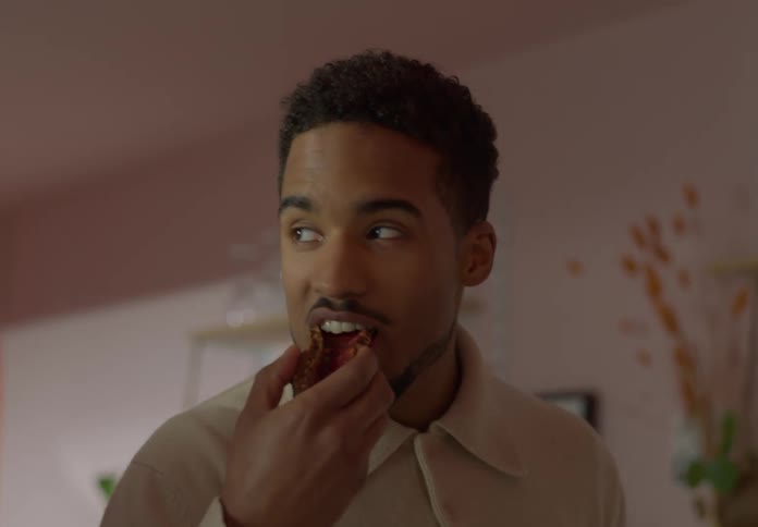 A man at home picks up a piece of bread and takes a bite and nudges his friend who is looking at the bread