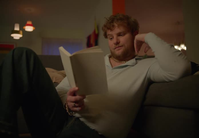 Man with beard and brown sweatshirt and jeans sits on couch reading a book in living room