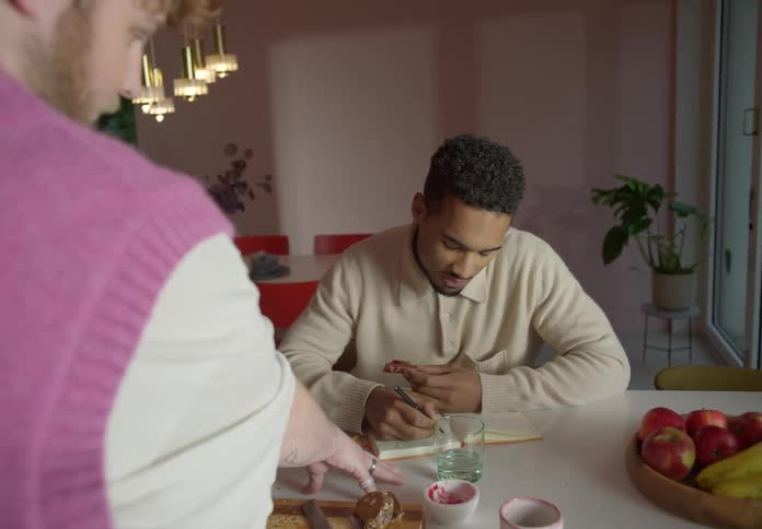 Man sits taking notes and eating while talking to another man in home