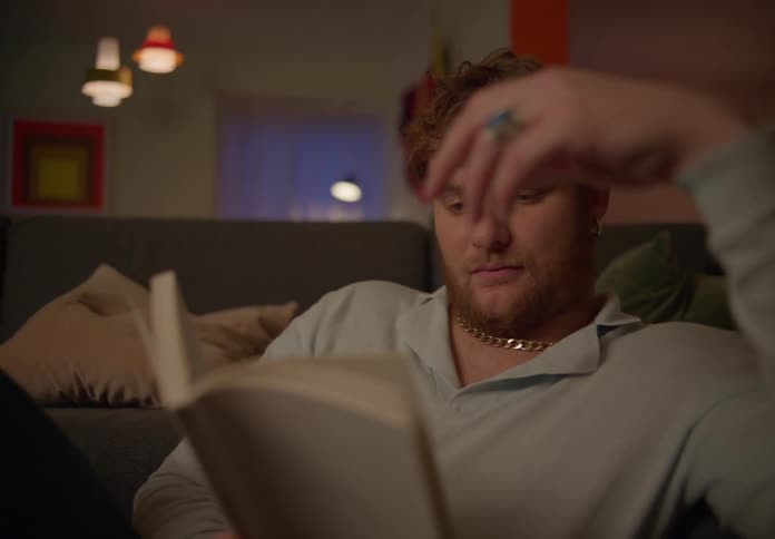A man is relaxing on the living room couch reading a book as he turns the page and laughs