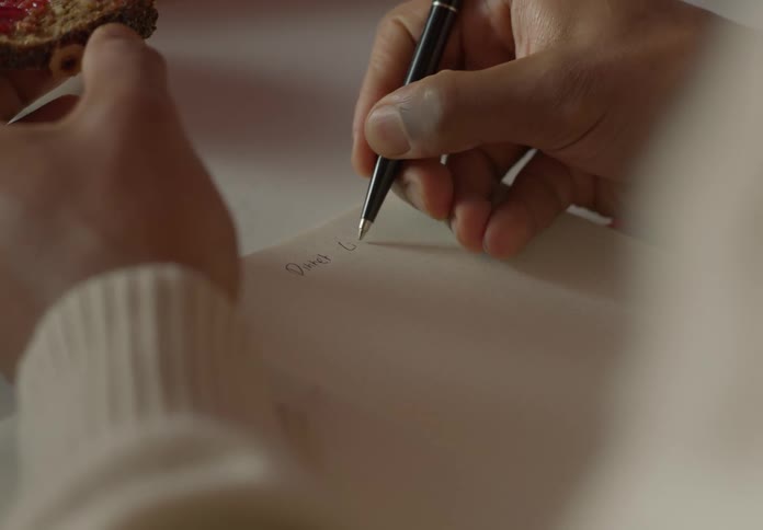 A man is holding a bagel in one hand as he writes on paper with the other hand