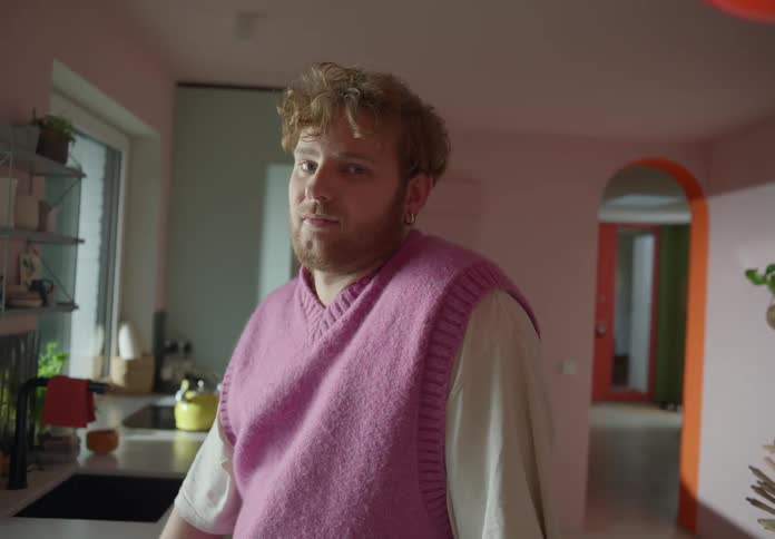 A bearded man wearing a purple vest looks out the window while standing in the kitchen then turns around and curiously looks at the camera