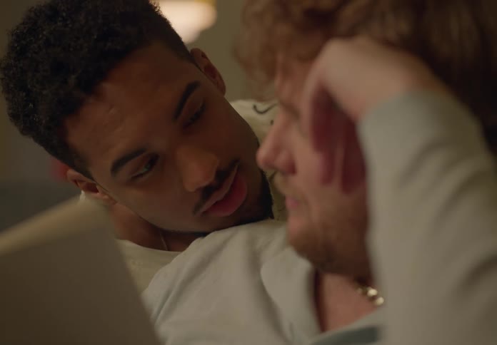 A man is sitting on the couch reading a book and his boyfriend is looking over his shoulder