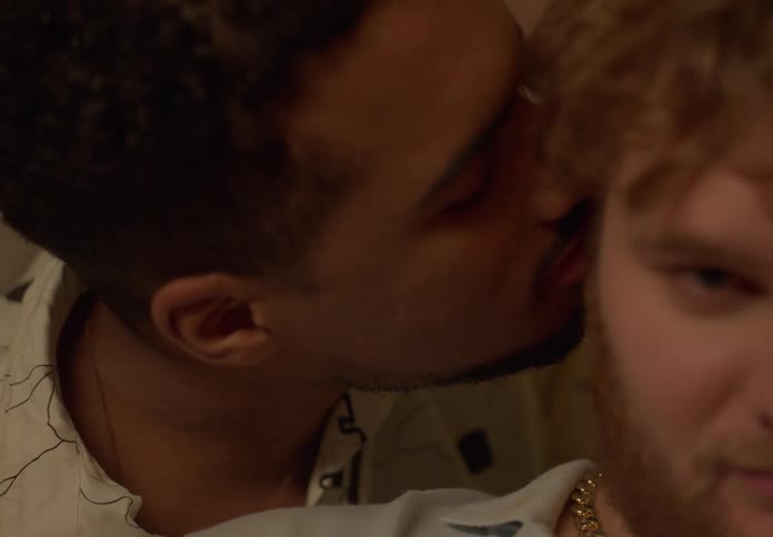 A man is standing behind his boyfriend as he leans over and kisses him on the neck and whispers in his ear