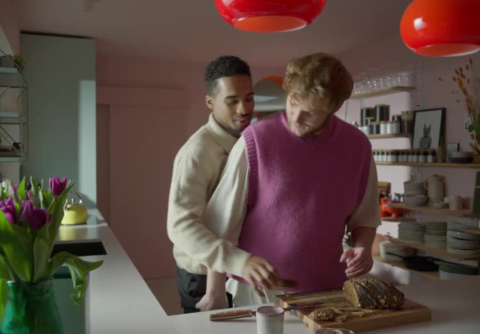 A man is standing over a table slicing bread and putting jelly on a slice of bread when his boyfriend walks in and kisses him on the cheek as he takes the slice and eats it