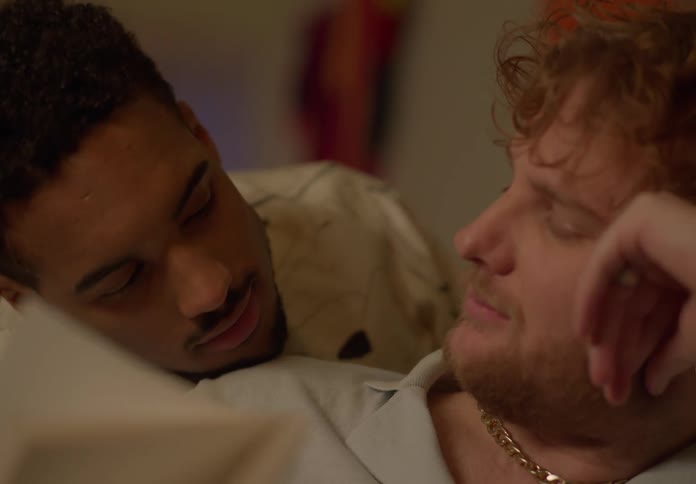 A man is sitting on the couch reading a book and his boyfriend is looking over his shoulder