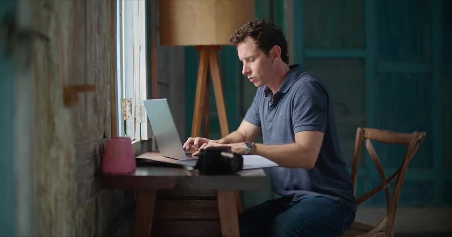 A man is sitting at a desk in front of a window looking through book and typing on a laptop computer
