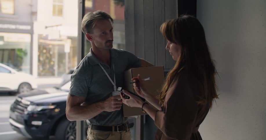 A delivery man is carrying boxes into a business as he hands the signature pad to a woman and she signs for the packages