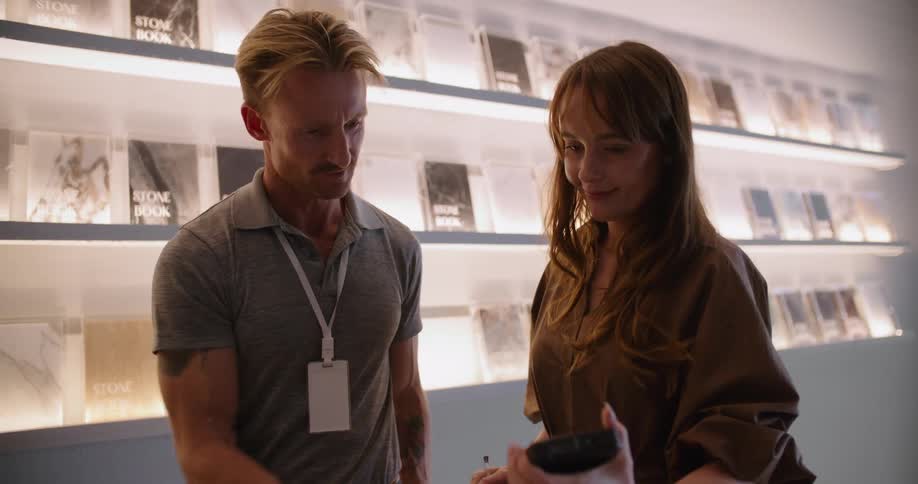 Blonde salesman with gray t shirt and brown pants takes electronic signature from woman with brown hair and dress at bookstore