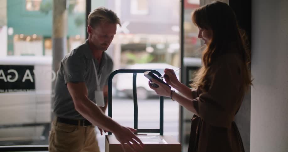 A delivery man wheels in a box to a business as he hands the signature pad to a woman and she signs for the package