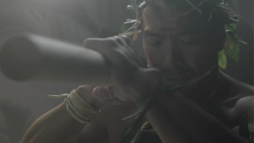 A man wearing a crown of leaves balances a long, wooden pole on his shoulders